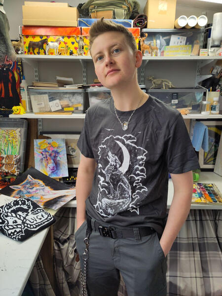 A photo of Hoggy wearing a grey t-shirt with his fox moon lino-cut design on the front. Hoggy has pale skin and blonde hair styled into a scruffy quiff. He looks to the left, behind him is his art studio.