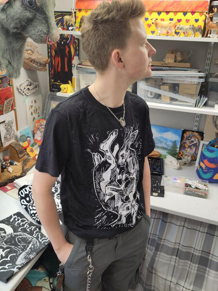A photo of Hoggy wearing a black t-shirt with his boxing hares lino-cut design on the front. Hoggy has pale skin and blonde hair styled into a scruffy quiff. He looks to the left, behind him is his art studio.
