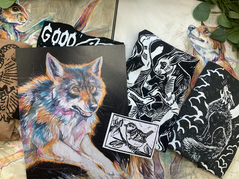 A collection of Sketchbuck's teemill products such as a digital print of a wolf, a digital lino-print of a wren, and t-shirt prints of boxing hares. Resting on two original canvas paintings.